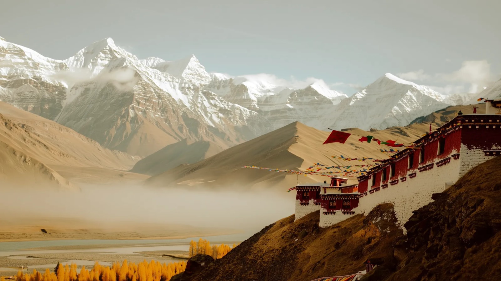 A whitewashed Tibetan monastery on a hillside below snow peaks, with mist in the valley